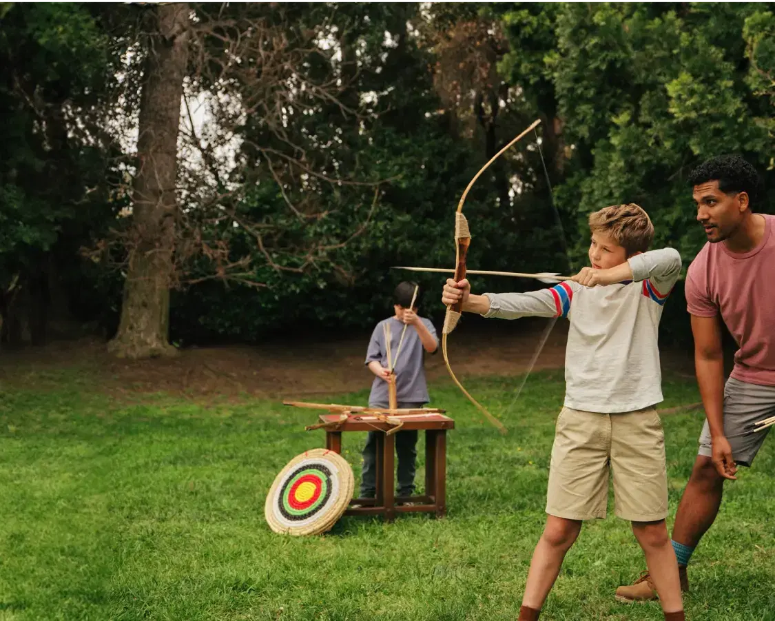A man patiently instructs a child in the art of archery, teaching them how to aim and shoot with a bow