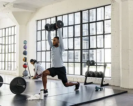 Man in a lunge lifting dumbbells in the gym with a woman in background